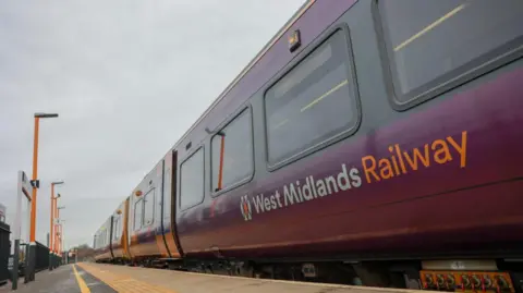 The side of train carriages pulled up on a platform. The words West Midlands Railway can be seen on one carriage