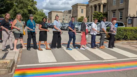 Staff standing in in a line across a zebra crossing with rainbow lines painted alongside it.