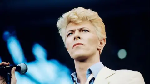 Getty Images David Bowie has bleached blond hair. He's wearing a white suit and a pale blue shirt and standing in front of a microphone