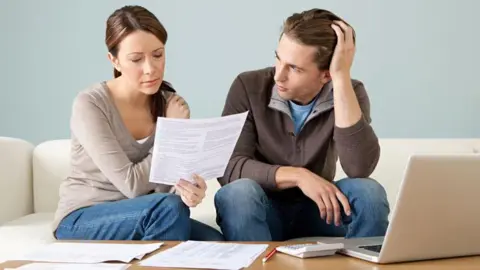 A posed image depicting a young couple going through their bills. On a table in front of them is paperwork and a laptop. 