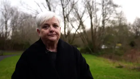 A woman with short white hair and a black jacket stands outside in a park