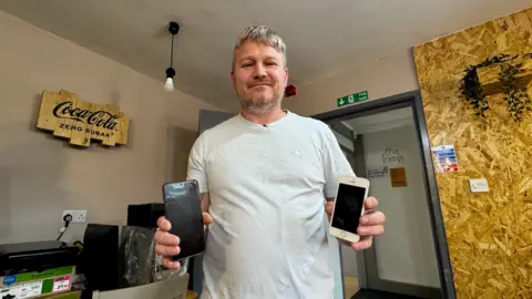 BBC A man with short light brown hair holds up two donated mobile phones. He is wearing a light blue-coloured T-shirt.