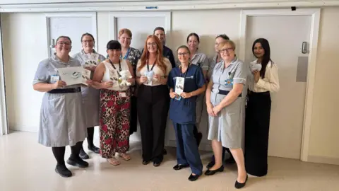 A group fo 11 women who are stood together in a white corridor, some of the women are in nurses uniforms and are holding certificates and teddy bears