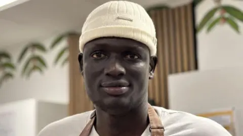 A man wearing a white woolie hat with a white T-shirt and brown apron looks into the camera.