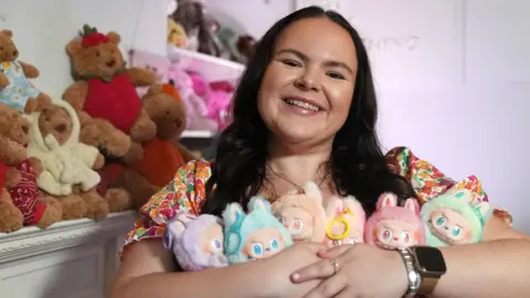 Meg, a young woman with long dark hair grins as the camera while holding six colourful Labubu dolls in different pastel colours. There are teddy bears behind her and a shelf of other colourful soft toys, which are slightly blurred in focus. 