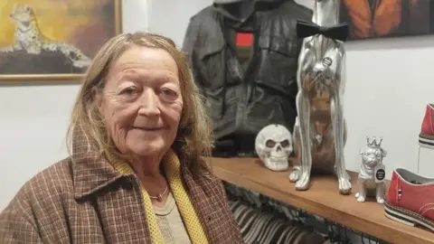Paul Burnell/BBC Nora Masefield has straw-coloured hair, a brown checked jacket with a yellow cardigan and caramel coloured sweater. She is standing in front of a shelf with red designer shoes, two metallic dogs and a fake skull.