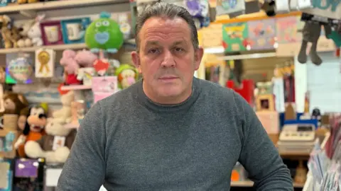 Simon Thake/BBC A heavily built man in a grey jumper stands stoney faced in front of a display of cards and soft toys.