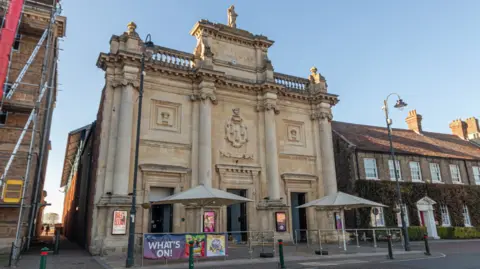 Getty Images The ornate frontage of King's Lynn Corn Exchange, faced in light-coloured stone with motifs showings sheaves of wheat. There are large cream-coloured parasols outside.