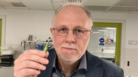 A man with a short grey beard and wearing glasses is looking at the camera. He's wearing a grey suit and holding a small black pellet in a pair of green tweezers. 