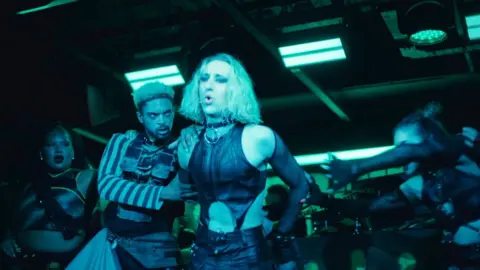 A group of dancers in outfits perform under vibrant teal lights, showcasing intense energy and movement.