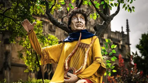 A puppet of Orpheus, taken in front of Gloucester Cathedral. He wears a yellow tunic and a blue scarf, and has brown curly hair. He holds a golden lyre in his lap with one hand, while the other is raised. 
