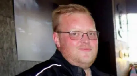 A man with short blond hair and stubble is pictured looking towards the camera while wearing a black motorcyclist jacket.
