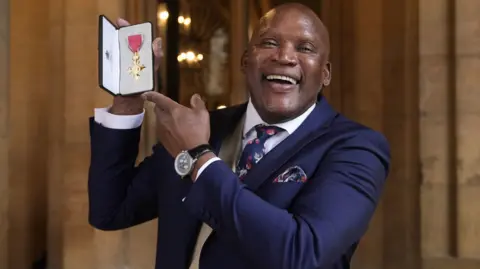 Devon Malcom smiles widely at the camera and is dressed in a navy suit with a matching patterned navy tie and handkerchief in his pocket. He is holding up his gold medal.