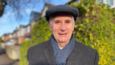 A man with a flat cap is pictured. He is wearing a blue and grey checked scarf and grey overcoat. Blurred houses can be seen in the background.