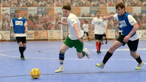 PA Media A futsal player wearing white dribbles past a rival wearing a blue bib. The match is being played on an indoor sports court with a blue surface.