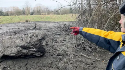 Gill in a yellow coat and warm hat points out the area she became stuck in.
You can see a large log to the left, and a dark-grey muddy area in front of her.