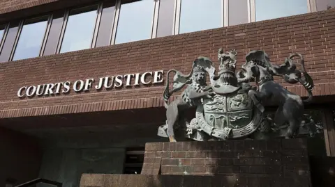 Getty Images Portsmouth Crown Court. A red brick building with a row of windows above a metal sign reading "Courts of Justice". A large metal statue of an insignia sits to the right of the sign. 