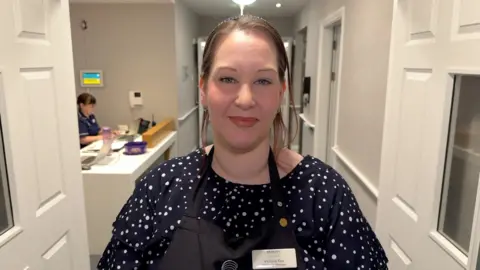 A woman stands in a corridor. She has straight brown hair. She is wearing a black and white polka dot shirt, a black apron, and a name badge that says "Victoria Key" on it.