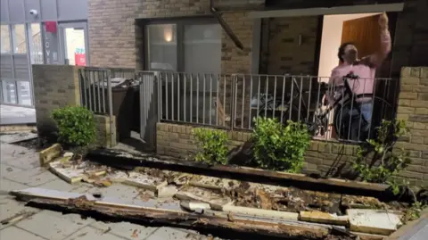 BBC Man pointing towards the collapsed balcony wreckage, with debris scattered across pavement outside a block of flats.