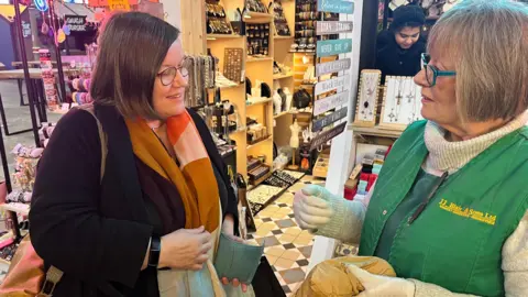 Dame Meg Hillier holding a purse and chatting with a greengrocer who is wearing a green work tunic and gloves. 