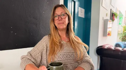A lady with long brunette hair, she is wearing glasses and a beige top. She is holding onto a green mug