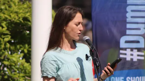 Karen Forsyth Hope stands outdoors at a microphone, reading from a mobile phone while speaking. She is wearing a light blue short‑sleeved top and has long brown hair. A dark banner with lettering is visible behind her, alongside greenery and a white column in bright daylight.