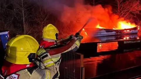 A boat on fire on a stretch of water is put out by firefighters 