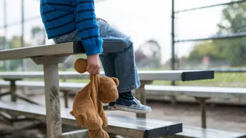 Getty Images A young boy sitting by himself on a stand of benches, holding a teddy bear