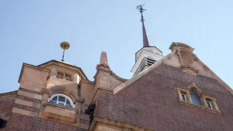 City of Wolverhampton Council A close-up view looking upward at the roofline of Wolverhampton Central Library, showing the newly replaced copper spire. The red brick and yellow terracotta dressings of the gable are sharp against a pale blue sky.