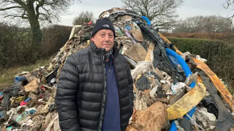 Councillor Phil Whitehouse, standing next to a large pile of waste material. He is wearing a black jacket over a blue jumper as well as a black woollen hat.