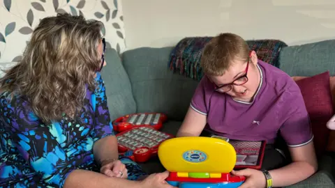 A woman in a blue and purple patterned blouse is say on a sofa with a teenage boy who is wearing a purple t-shirt. The boy is holding a brightly-coloured toy and there are others on the sofa next to him.