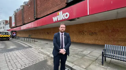  In the middle of the photo is Alex McIntyre, arms crossed wearing a suit and tie. Behind him is a boarded-up Wilko store with red signage and logo directly above the MPs head. Above the signage and shop livery is a red brick front. Below is the boarded up frontage that was in the previous photo just from an angle.
