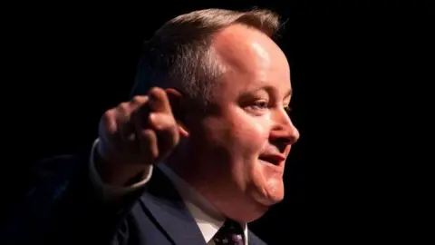 Getty Images Darren Millar is pointing towards the camera while looking away at a right angle towards an audience. He is wearing a suit, tie and shirt, and is stood before a dark background.