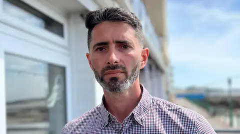 Florin Beclenas on the promenade. He has a dark speckled bears and short hair. he is wearing a checkered shirt.