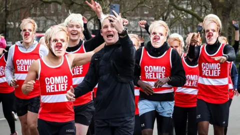 Getty Images Jamie Laing dengan pelari mengenakan topeng laing pada pagi hari pertama di 2025 komiknya tantangan bantuan di Hyde Park pada 17 Maret 2025 di London, Inggris. Pelari mengenakan rompi merah dengan logo relief komik yang ditulis dengan huruf putih tebal, dan beberapa mengenakan wig pirang