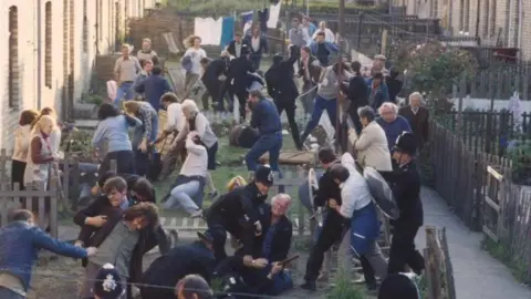 A screenshot from the Our Friends in the North scene where there is a violent disturbance in a pit village. It looks to be a mass fight with the police in the front gardens of terraced houses. Fences have been knocked over and there are multiple people on the ground. There are people of all ages there.