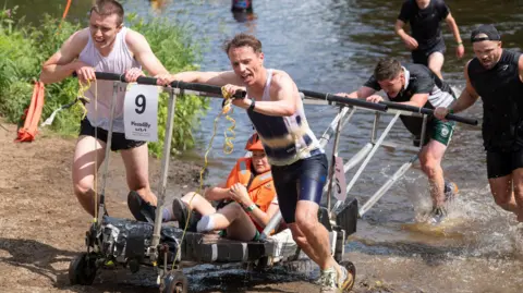 A team struggles through the River Nidd with their bed