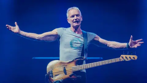 Getty Images Sting is pictured performing on stage during a show in Milan, Italy. He has his arms stretched out wide while he sings and a guitar hangs from his body.