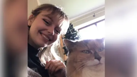 Kat Nellist Kat Nellist in a selfie with a light brown cat. The catl has a white neck and green eyes and is looking to the right of the camera. Kat is smiling as she takes the photo