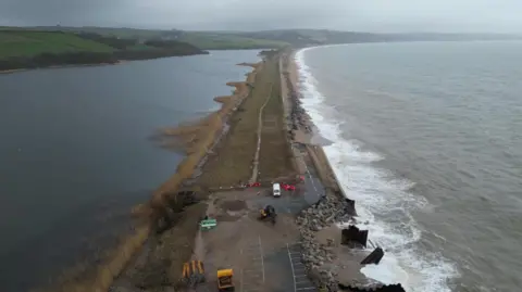 A drone view of the A379 Slapton Line between Torcross and Slapton. It shows the sea on one side and a lake on the other side. The road is damaged in two parts where maintenance vans can be seen.