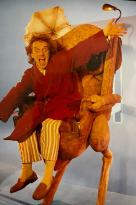 Bob Baldwin Rik Mayall wearing stripy pyjama trsouers, slippers and a red dressing gown. He is sitting in a strange chair which has the legs of a bird. His arms are spread and his mouth is wide open as he looks at the camera.