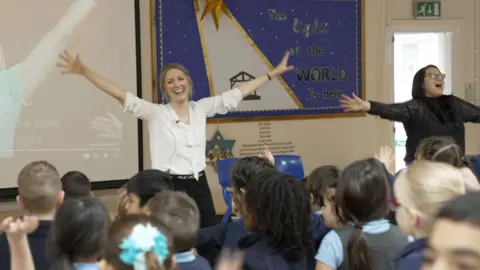 Head teacher Mrs Booth is standing in an assembly hall in front of the whole school with her arms outstretched with another teacher, they're mid-song. 