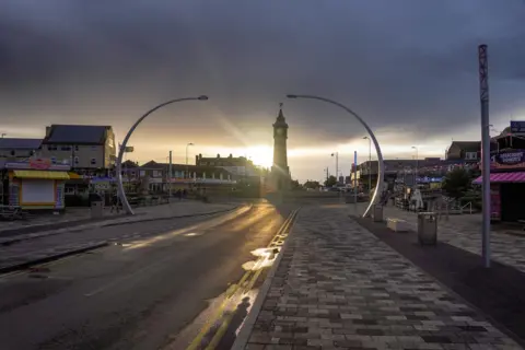 Benzla A view of Skegness taken from Tower Esplanade looking towards the clock tower. The sun is setting behind the clock tower. Heavy grey clouds fill the sky. Curved streetlights are framing the clock tower. It has recently rained and there are puddles in the road. 