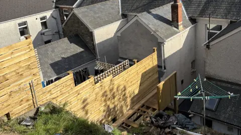 A timber raised platform connecting a brick wall and a grey brick house. 