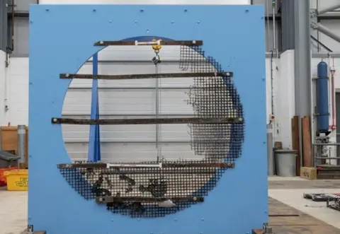 HSE Industrial test setup featuring a large circular panel mounted on a blue frame, with metal reinforcement bars and partially damaged wire mesh across the surface, inside a workshop with tools, pipes, and equipment in the background.