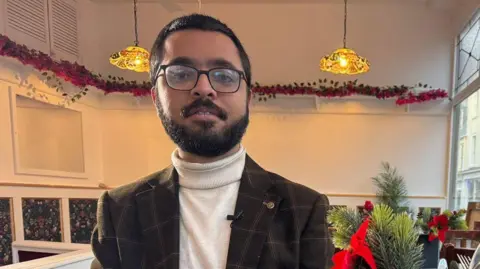 Muhammad sits in his restaurant; the decorated walls are visible in the background. He is wearing a white polo neck and a brown plaid jacket. He has dark brown hair and full beard. He is wearing glasses
