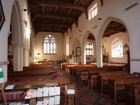 Andrew Hill/Geograph The interior of All Saints Church, Holbrook. The nave has white-washed arches along its sides. The altar is at the far end with an arched window behind it. The ceiling has wooden beams.