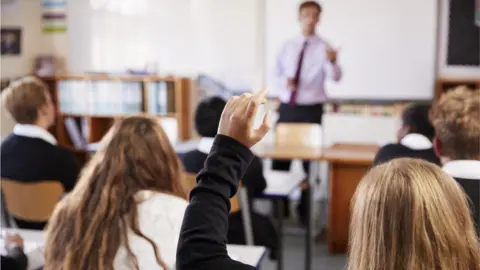 Getty Images A teacher teaching pupils in a classroom in front of bookshelves and a whiteboard. In the foreground, a girl has her hand up 