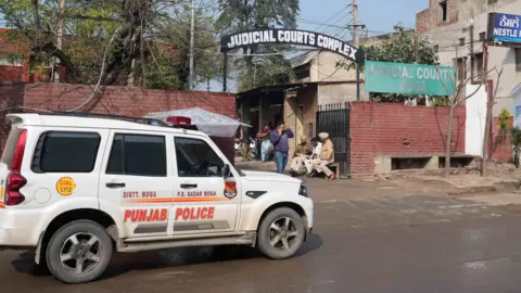 Adnan Bhat A Punjab Police car parked outside the entrance to the Judicial Courts Complex in Moga. There are black gates to the complex, between red brick walls, and a black sign above the entrance road. Three men are sitting on seats at the entrance and a man is talking on a mobile phone.