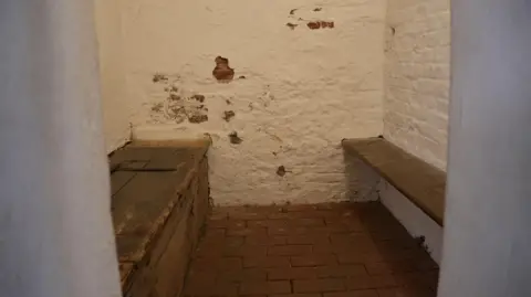 Karl Bird / BBC The Pilgrims' cells at Boston Guildhall features two wooden benches, a red brick flooring and walls painted white. 
